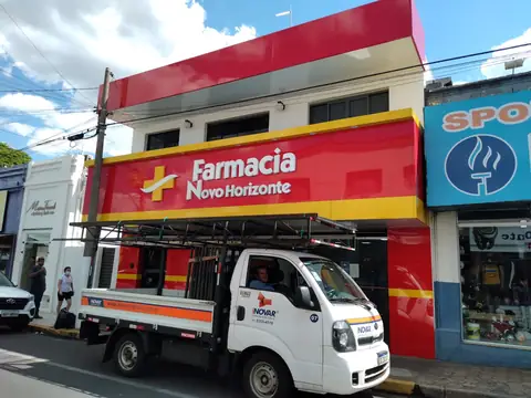 Fachada em ACM vermelho e amarelo da Farmácia Novo Horizonte, com caminhão com carroceria à frente