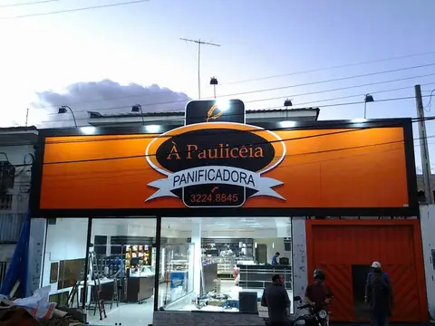 Fachada em ACM laranja com letreiro luminoso da À Paulicéia Panificadora, iluminada por refletores