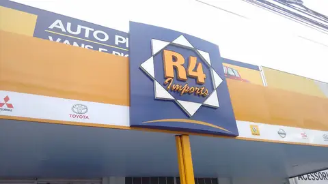 Placa em poste com logo losango da R4 Imports sobre fachada amarela e branca de autopeças