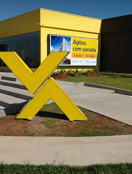 Escultura grande em forma de X amarelo diante de fachada amarela, em jardim com escada