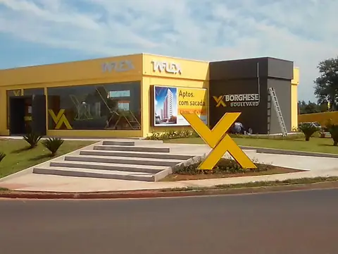 Fachada amarela com letras caixa e grande X amarelo à frente, com escada de acesso e gramado