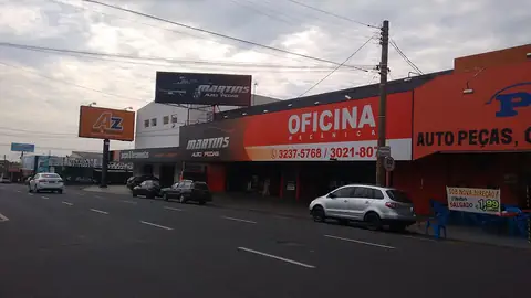 Fachada de autopeças com faixa laranja e letreiros impressos em avenida, ao entardecer
