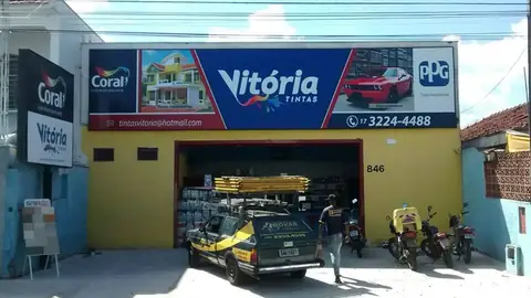 Painel impresso azul da Vitória Tintas com logos Coral e PPG sobre fachada amarela