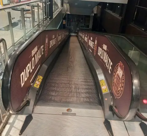 Esteira rolante de shopping com adesivos impressos em vinho nos corrimãos, texto Duplo Malte