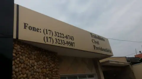 Marquise curva em ACM bege com telefones e áreas de atuação em letras pretas, base de pedra