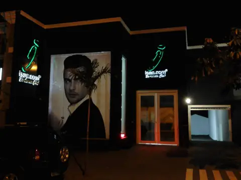 Fachada noturna com logotipos neon verdes isac.com e painel com retrato, vista à noite