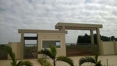 Pórtico de entrada de condomínio com pilares bege, guarita e palmeiras, céu nublado