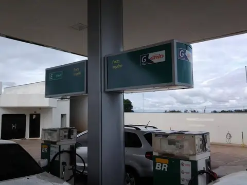 Posto de combustível com totens e placas iluminadas verdes sob a cobertura, bombas ao fundo