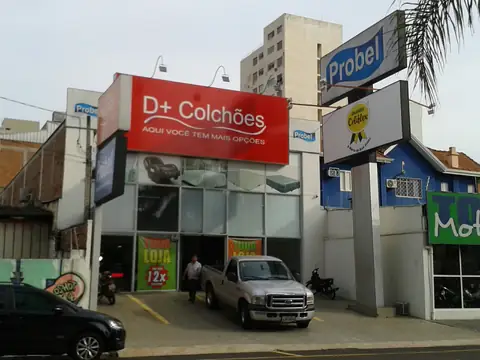 Painel luminoso vermelho da D+ Colchões na fachada da loja, com placas em poste ao lado