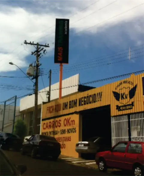 Fachada amarela de loja de carros com totem luminoso vertical e faixa de letras vermelhas