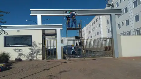 Pórtico de condomínio em ACM cinza com instaladores em andaimes e prédios ao fundo