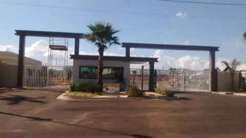 Dois pórticos escuros sobre a entrada de condomínio, com guarita e palmeira