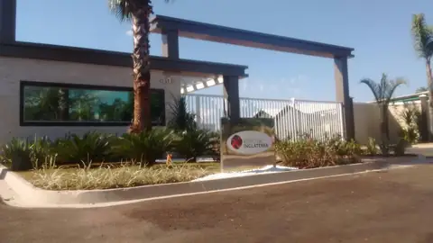 Entrada de condomínio com pórtico escuro, placa em canteiro e portão branco