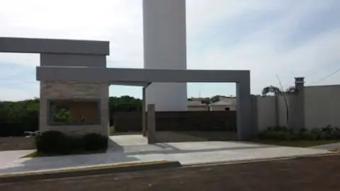 Pórtico em ACM cinza sobre guarita de pedra, com torre alta ao fundo