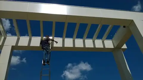 Instalador em escada sob pergolado de ACM branco, visto de baixo contra o céu azul