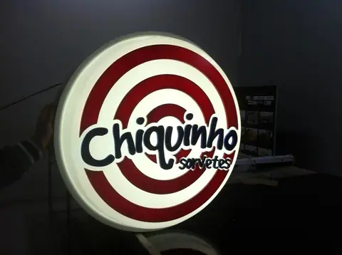 Luminoso circular com listras vermelhas e logo Chiquinho Sorvetes aceso em ambiente escuro