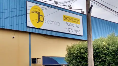 Painel branco com logo amarelo da Jb Bechara Móveis sobre fachada azul e spots de iluminação