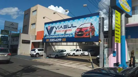 Grande painel azul da Viasul Auto Locadora com carro vermelho sobre a calçada