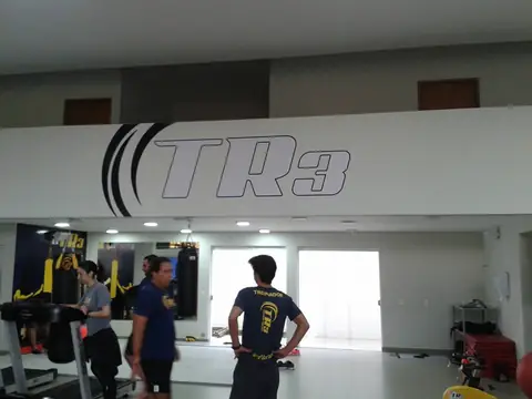 Logotipo TR3 em grande adesivo sobre parede branca de academia, com alunos em treino