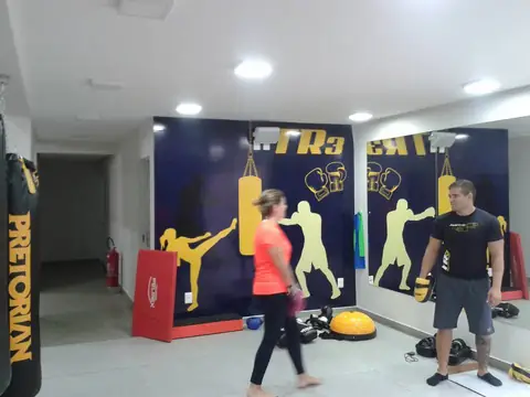 Academia com painel azul e amarelo de lutadores e logo TR3, pessoas treinando