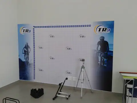 Backdrop impresso com ciclistas e logos TR3 em parede branca, com tripé ao lado